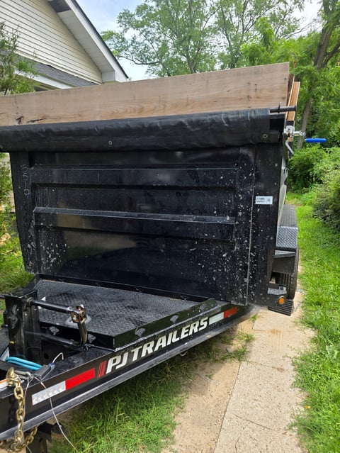 Black PJ Trailers dump trailer with open bed parked on residential driveway with trees and house in background