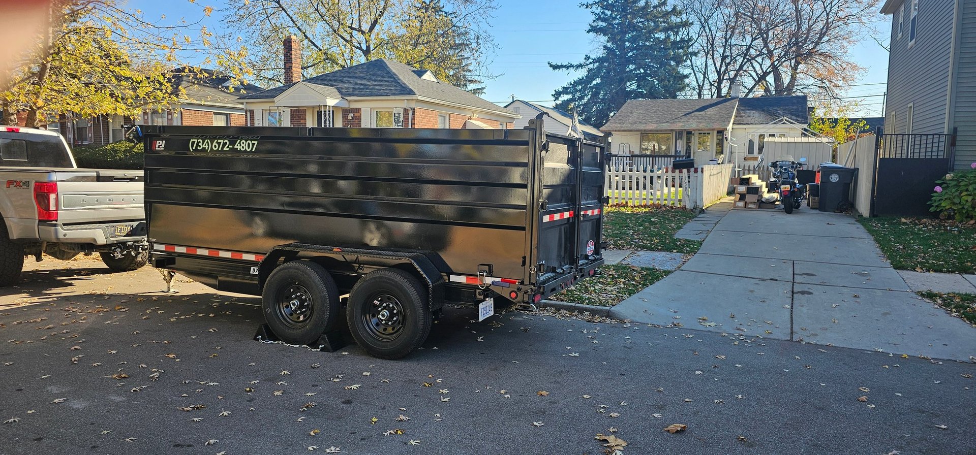 Black dump trailer parked on residential driveway with autumn trees and houses in background